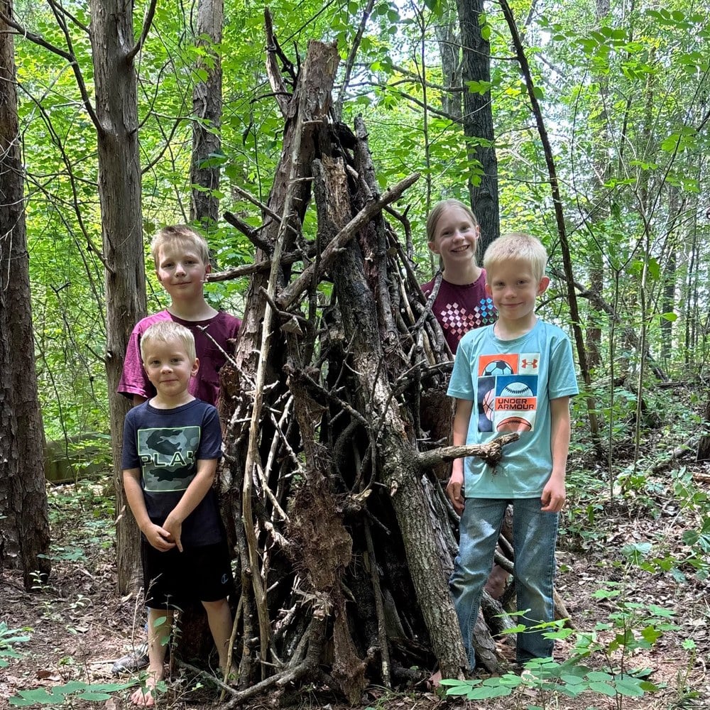 Four children standing beside the stick fort they built in the woods