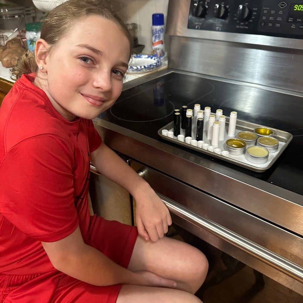 A girl crouched by the oven beside a tray of lip balm tubes and tins