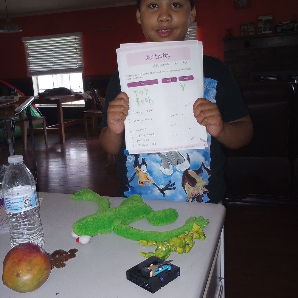 A boy holding up the needs versus wants worksheet he filled in